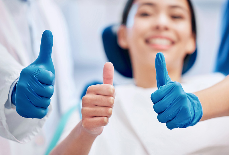 Image Two thumbs up with blue medical gloves, a smiling person in the background wearing medical attire.